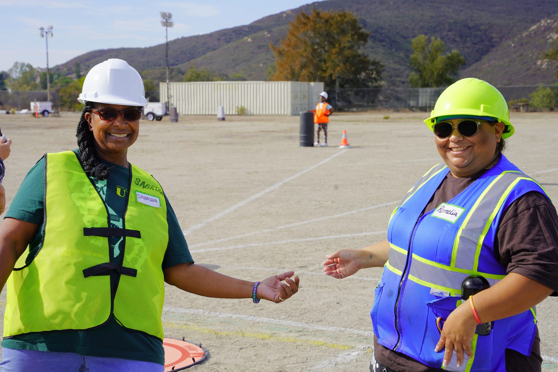 Two smiling drone technology students in hard hats and safety vests on an outdoor flight training field